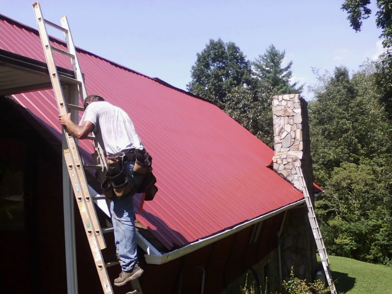 Roofer on a ladder beside a red metal roof and stone chimney — Soffit & Fascia Repair by Roofing Brothers in Norwell