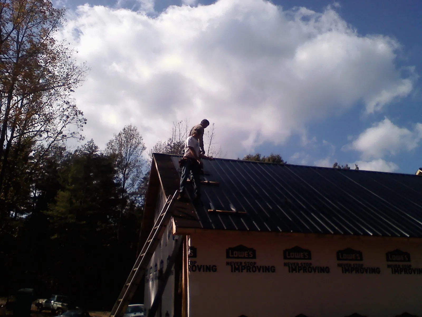 Roofer fastening dark metal panels high against a cloudy sky, from a Roof Inspection job in Norwell